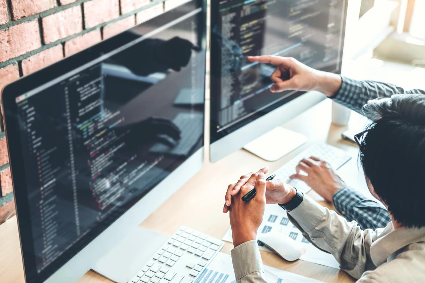 Person pointing at code on dual monitor setup while working at desk with brick wall background.