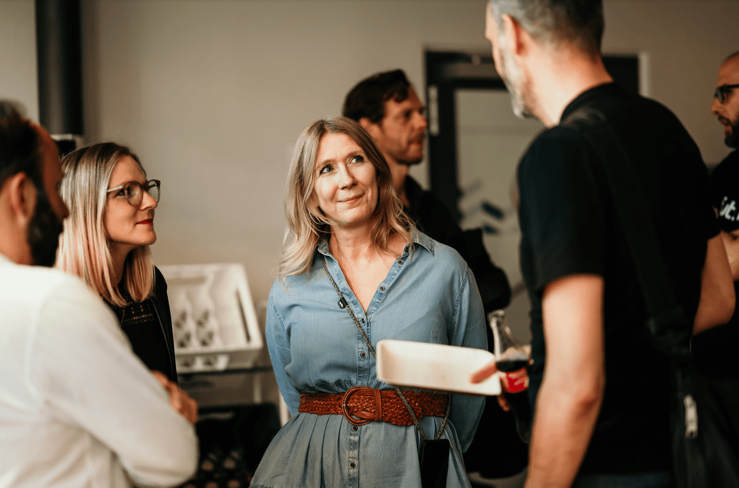 A woman in denim dress with brown belt engaged in conversation at a networking event or informal gathering.