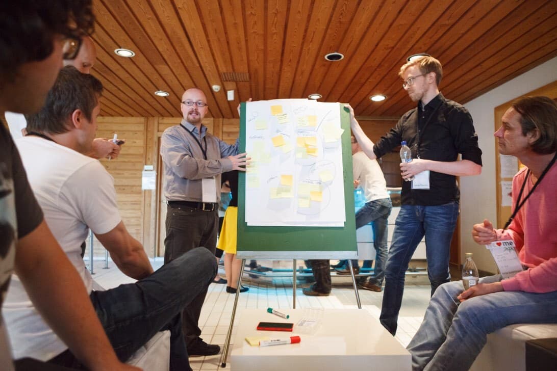 Team workshop with two people presenting a board covered with sticky notes to colleagues in a wood-panelled meeting room.