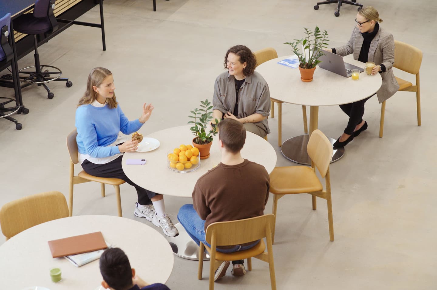 People conversing at round tables in a modern office space with wooden chairs, potted plants and a bowl of oranges.